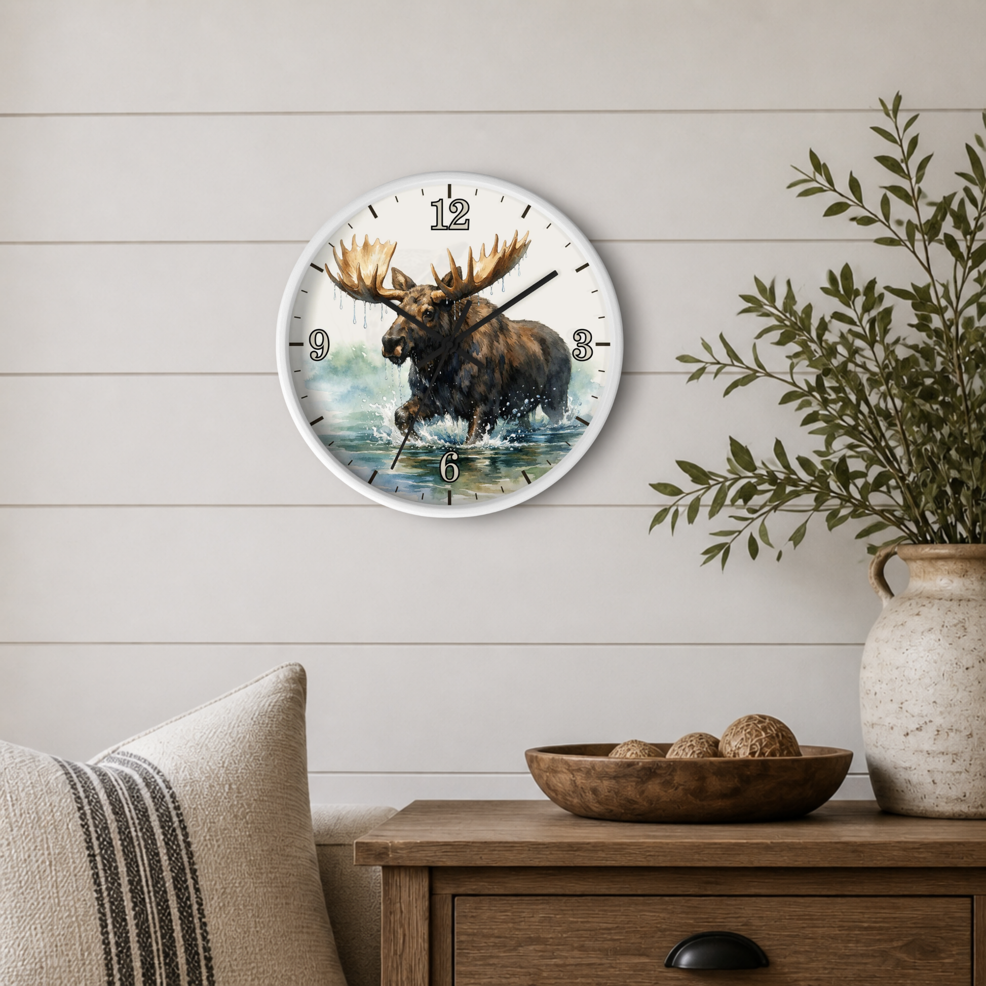 A round wall clock with a moose illustration in the center, mounted on a white shiplap wall above a wooden side table with a bowl and a potted plant.