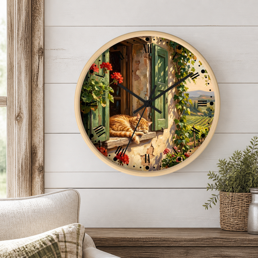 Round wall clock with a scenic print of a cat sleeping on a windowsill, surrounded by green shutters, red flowers, and vineyard views. The clock is mounted on a white wood-paneled wall above a wooden shelf with a plant and cushion.