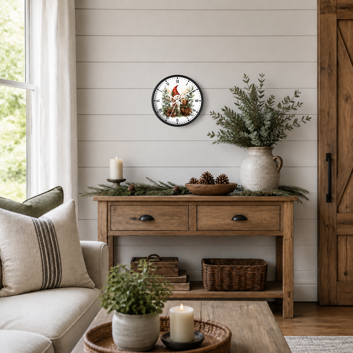 A round wall clock featuring a festive snowman and bear illustration, styled in a cozy, rustic living room setting above a wooden console table with holiday greenery and candles.