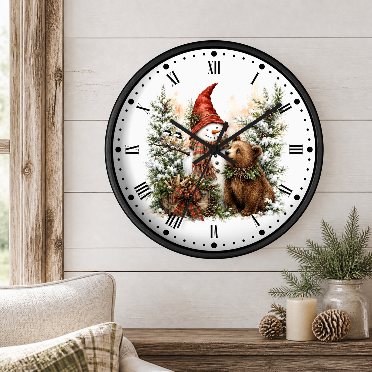 A round wall clock with a black frame and a winter woodland bear and snowman illustration on the face, hanging on a light wood paneled wall above a table with candles and pinecones.