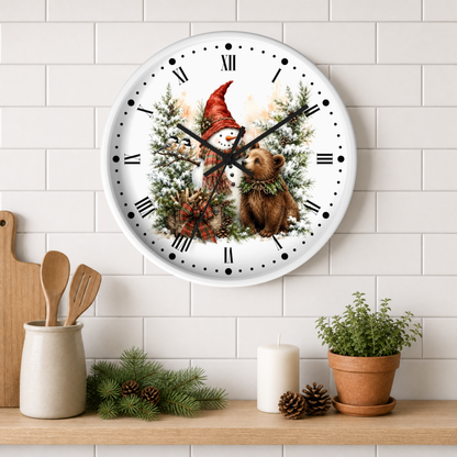 A round wall clock with a white face and black Roman numerals, featuring a festive illustration of a snowman in a red hat and a brown bear among pine trees. The clock is mounted on a white subway tile wall above a wooden shelf styled with kitchen utensils, candles, pinecones, and a potted plant.