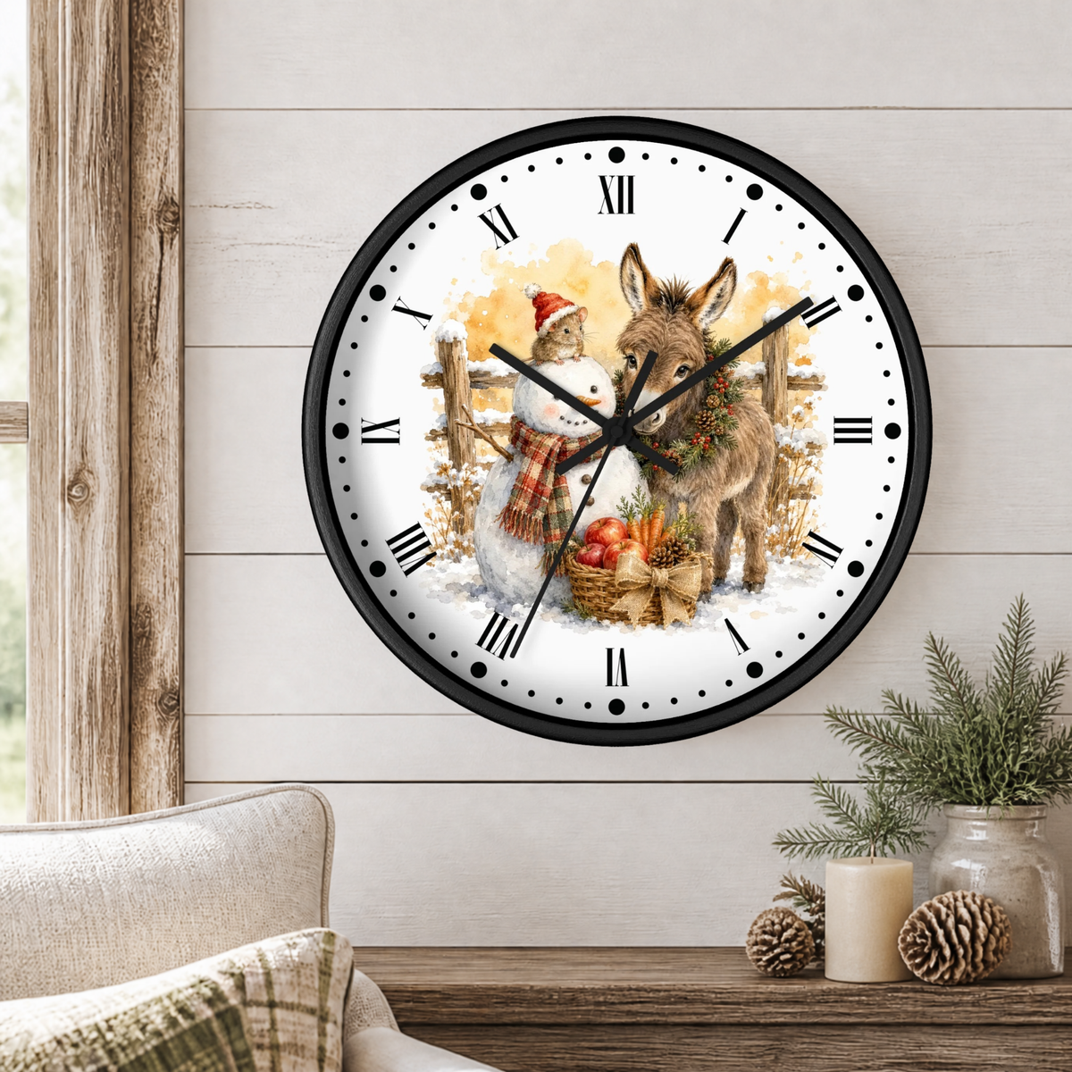 A round wall clock with a black frame and a festive winter illustration of a snowman and a donkey, displayed on a light wood paneled wall above a rustic table with candles and pinecones.