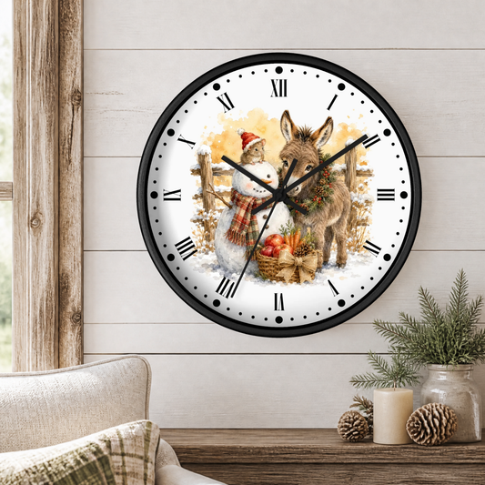 A round wall clock with a black frame and a festive winter illustration of a snowman and a donkey, displayed on a light wood paneled wall above a rustic table with candles and pinecones.