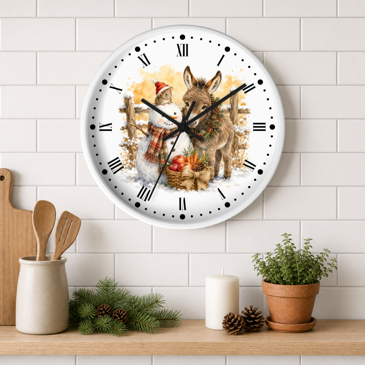 A round wall clock with a white face and black Roman numerals, featuring a festive illustration of a donkey and a snowman with pumpkins. The clock is mounted on a white subway tile wall above a wooden shelf styled with kitchen utensils, pine branches, a candle, and a potted plant.
