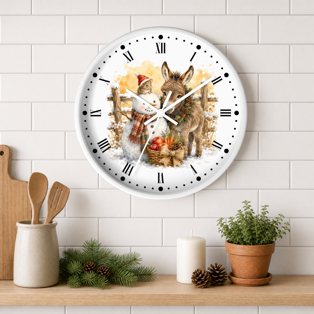 A round wall clock with a white face and black Roman numerals, featuring a festive illustration of a donkey and a snowman. The clock is mounted on a white subway tile wall above a wooden shelf styled with kitchen utensils, pine branches, a candle, and a potted plant.