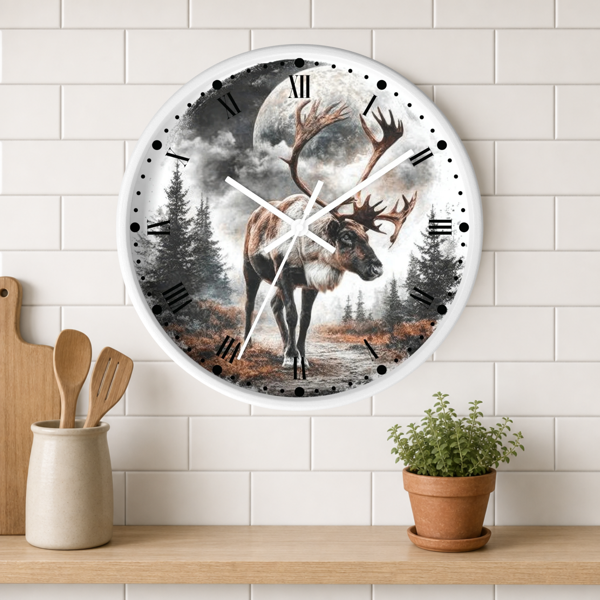 A round wall clock with a white frame and Roman numerals, featuring an illustration of a large deer with antlers standing in a misty forest under a full moon. The clock is mounted on a white subway tile wall above a wooden shelf with kitchen utensils and a potted plant.