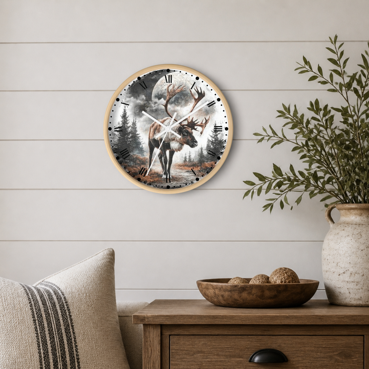 A round wall clock with a light wood frame hangs on a white shiplap wall above a wooden side table. The clock face features a moody forest scene with a reindeer, moon, and smoke, styled in a cozy home interior with a vase of greenery and a striped pillow.