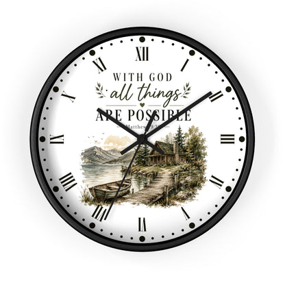 A round wall clock with a black frame and a white face featuring the text 'WITH GOD all things ARE POSSIBLE' in black and gold script, along with a scenic illustration of a lakeside cabin and mountains. The clock is shown against a plain white background.