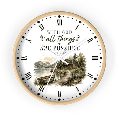 A round wall clock with a light wood frame and a white face featuring the text 'WITH GOD all things ARE POSSIBLE Matthew 19:26' above a scenic illustration of a cabin by a lake. The clock uses black Roman numerals and hands on a clean white background.