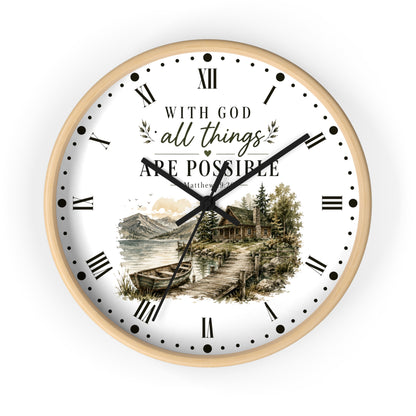 A round wall clock with a light wood frame and a white face. The clock face features black Roman numerals, black hands, and a scenic illustration of a lakeside cabin with mountains and trees.
