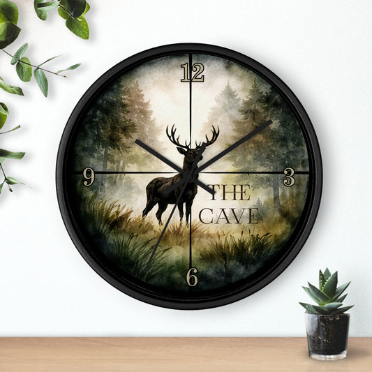 A round black wall clock with a forest scene and a deer silhouette, featuring the words 'THE CAVE' and stylized numbers, shown mounted on a white wall above a wooden surface with small plants.