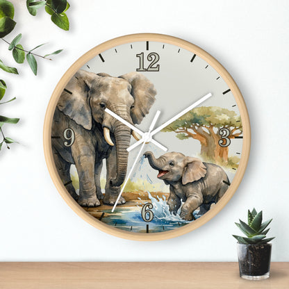 A round wall clock with a light wood frame and a printed safari scene featuring an adult and baby elephant, water, and trees. The clock is mounted on a white wall above a wooden surface, with decorative green leaves and a small potted succulent nearby.