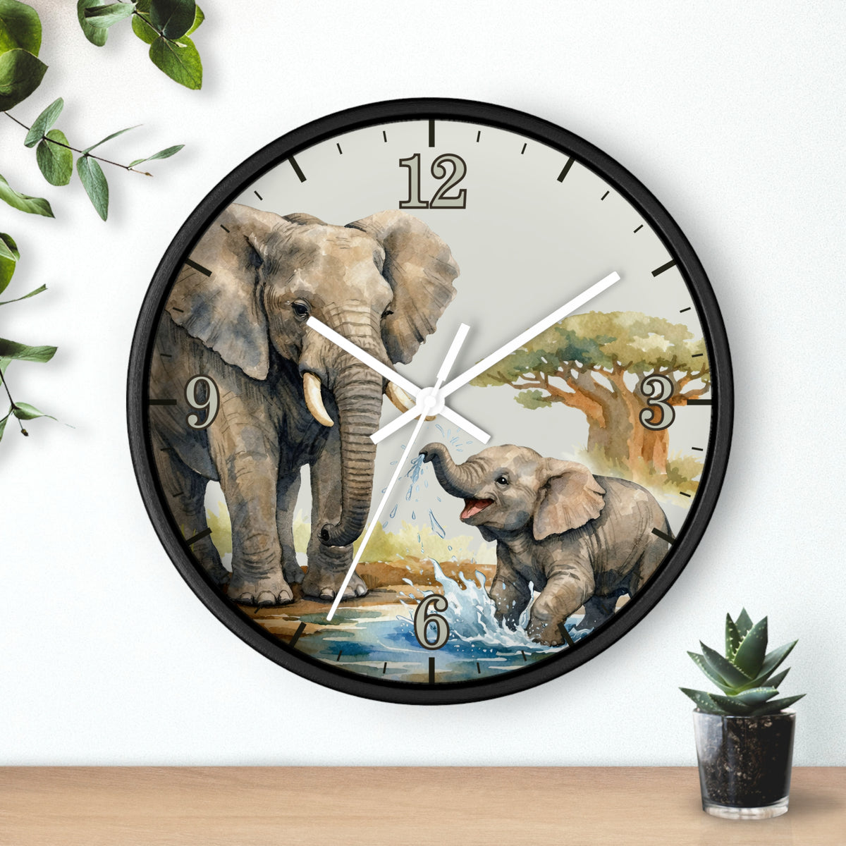 Round wall clock with a black frame and a printed scene of an adult and baby elephant at a watering hole, shown hanging on a white wall above a wooden surface with small plants.
