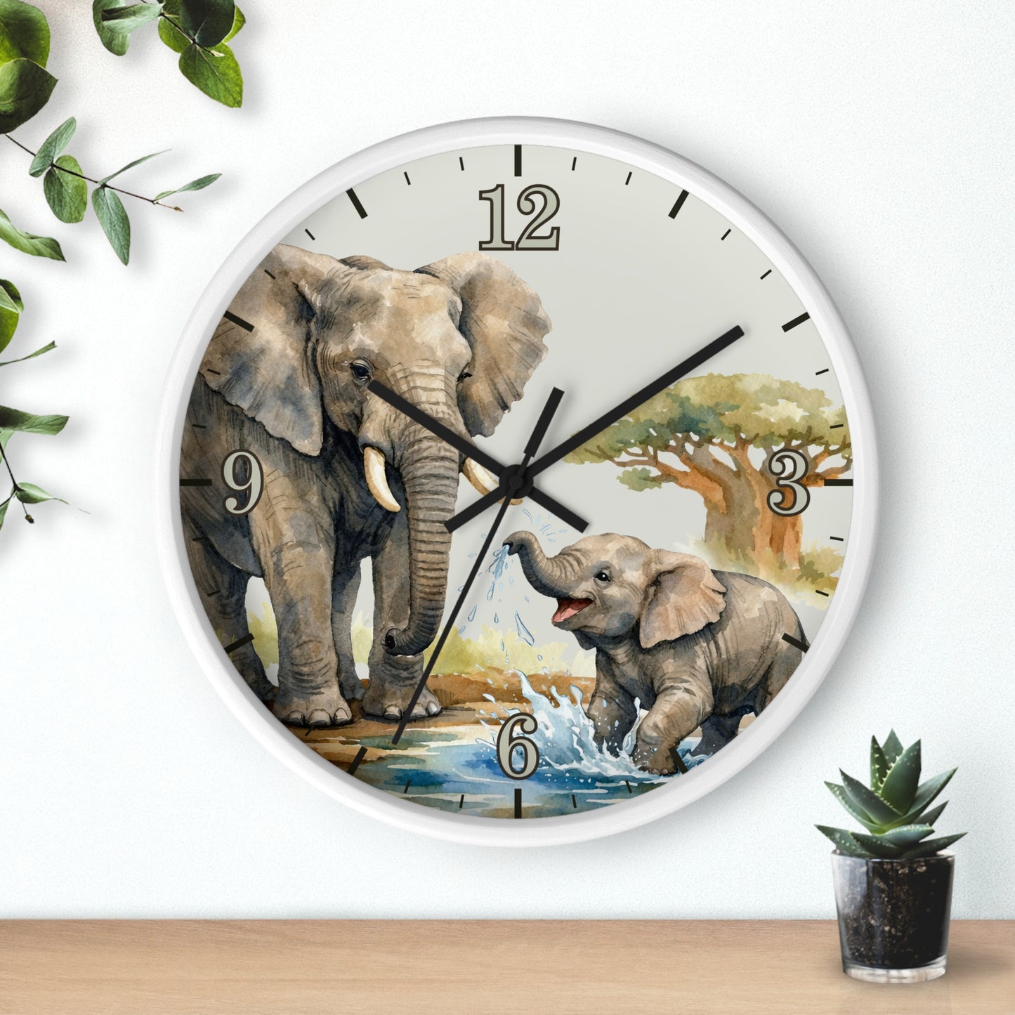 A round wall clock with a white frame and a printed illustration of an adult and baby elephant at a watering hole. The clock is mounted on a white wall above a light wood surface, with decorative green leaves and a small potted succulent nearby.
