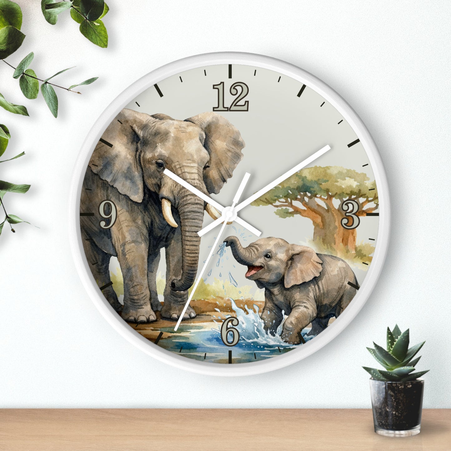 Round wall clock with a white frame and a printed scene of an adult and baby elephant at a watering hole, shown hanging on a white wall above a wooden surface with small plants nearby.