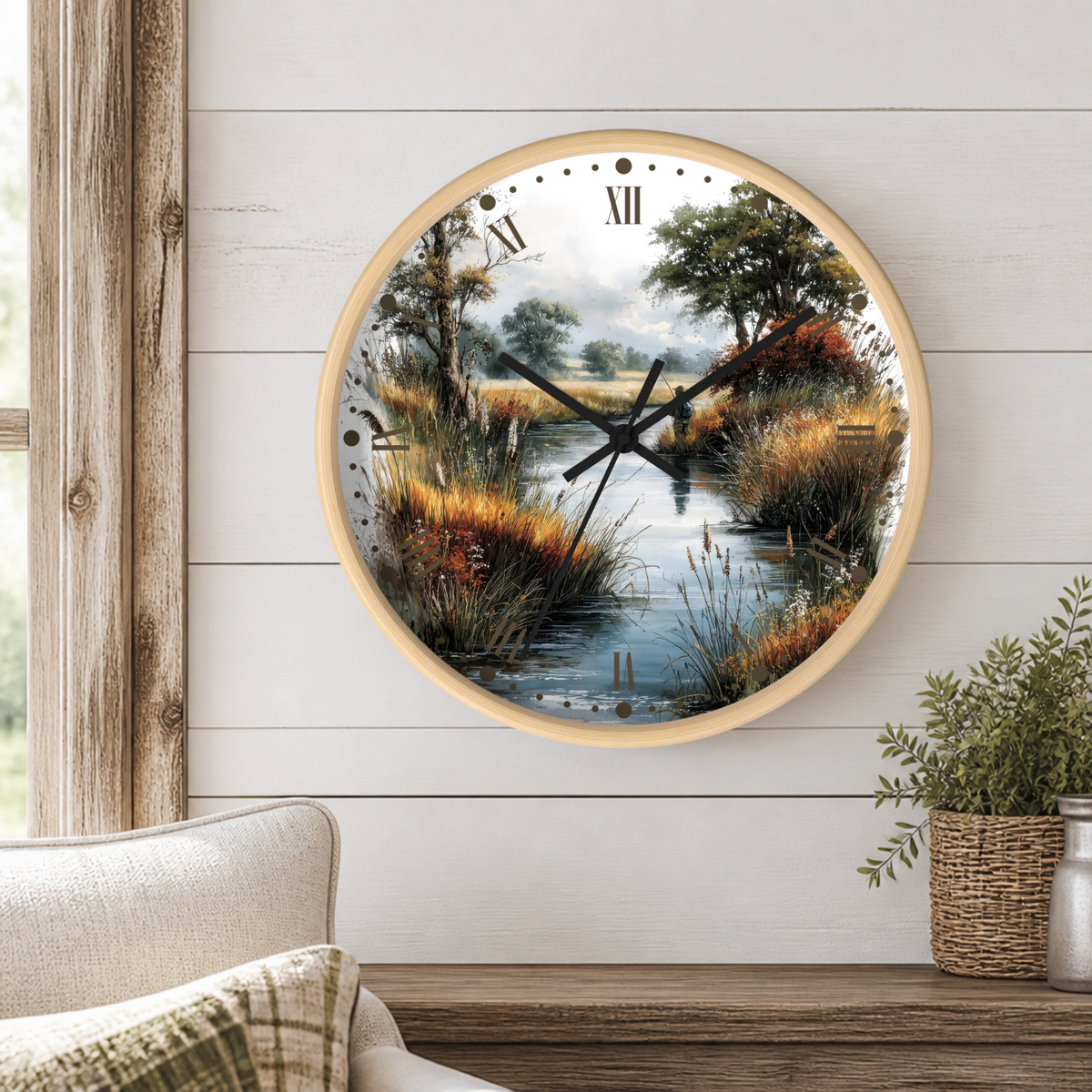 A round wall clock with a light wood frame and a scenic landscape print of a marsh or river landscape with trees, reeds, and water. The clock is mounted on a white shiplap wall above a wooden mantel with a small potted plant.