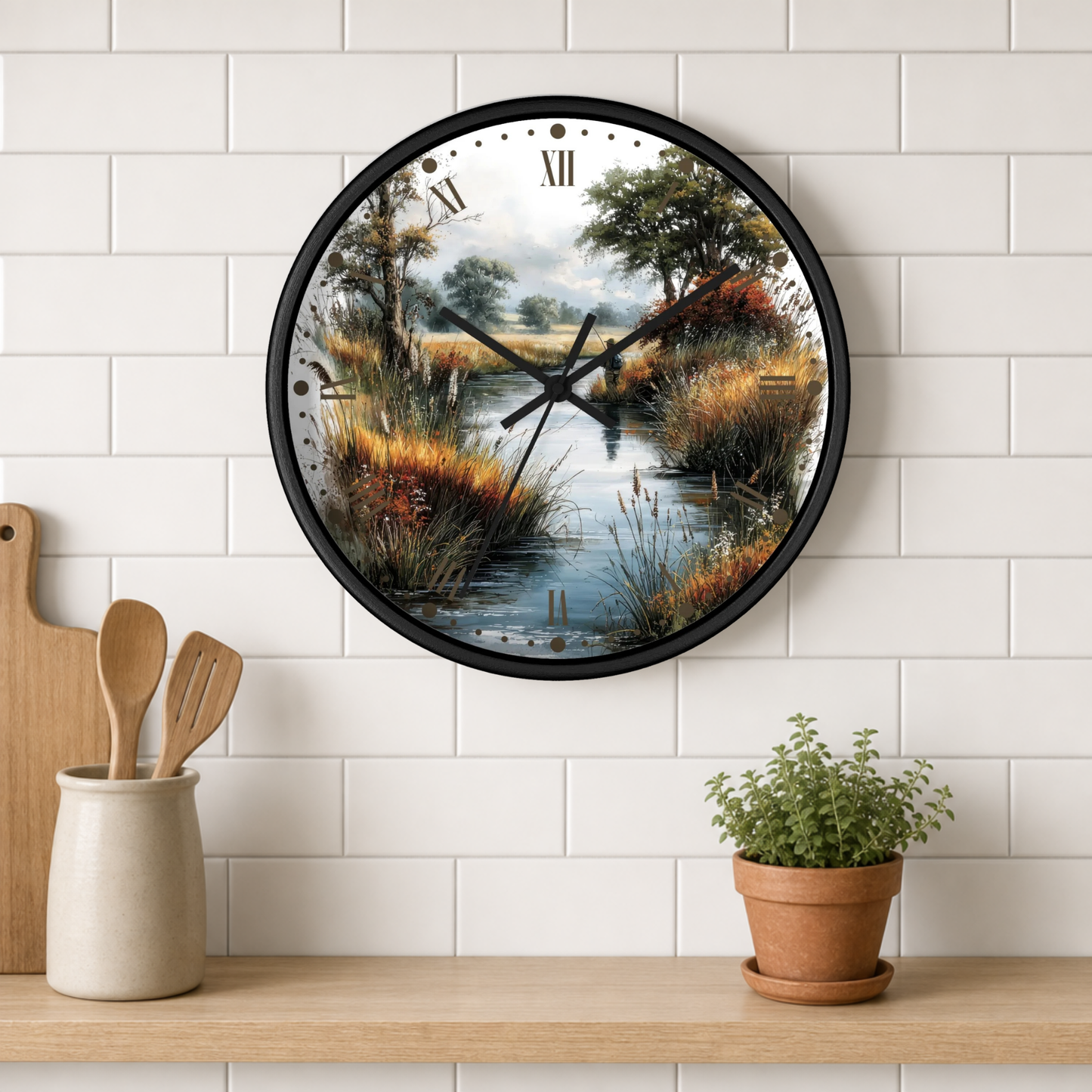 A round wall clock with a black frame and a scenic landscape print of a river crossing by a marsh, mounted on a white subway tile wall above a wooden shelf with kitchen utensils and a potted plant.