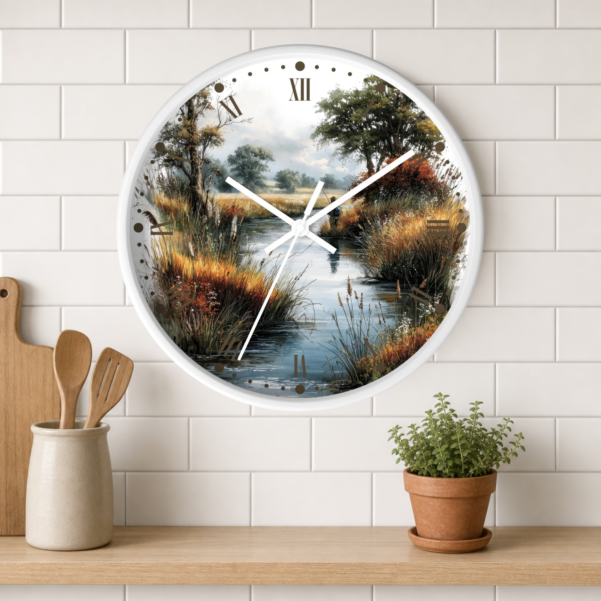 A round wall clock with a white frame and a scenic landscape print of a marsh or creek, styled on a white subway tile wall above a wooden shelf with a small potted plant and wooden utensils.