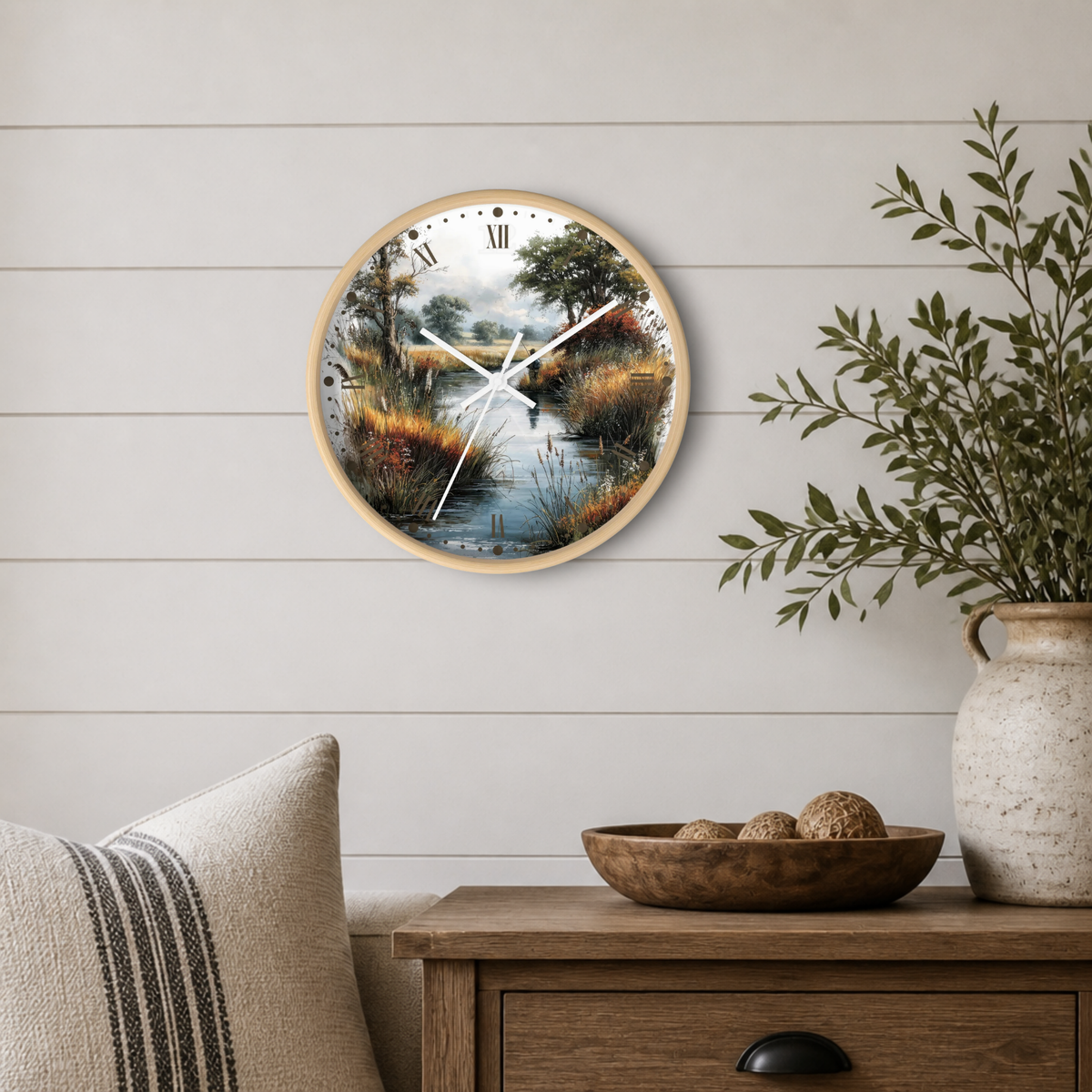 A round wall clock with a light wood frame and a scenic landscape print of a river marsh, mounted on a white shiplap wall above a wooden side table with a plant and decorative bowl.