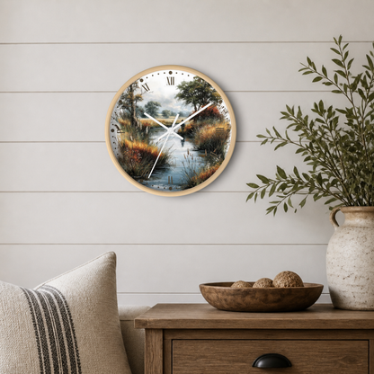 A round wall clock with a light wood frame and a scenic landscape print of a river marsh, mounted on a white shiplap wall above a wooden side table with a plant and decorative bowl.
