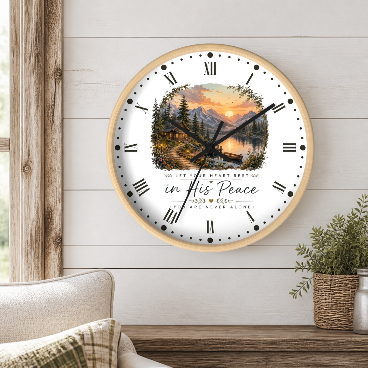 A round wall clock with a light wood frame hangs on a white shiplap wall above a wooden table. The clock face features a scenic landscape illustration of a mountain lake at sunset, Roman numerals, black hands, and the text 'in His Peace' and 'YOU ARE NEVER ALONE.
