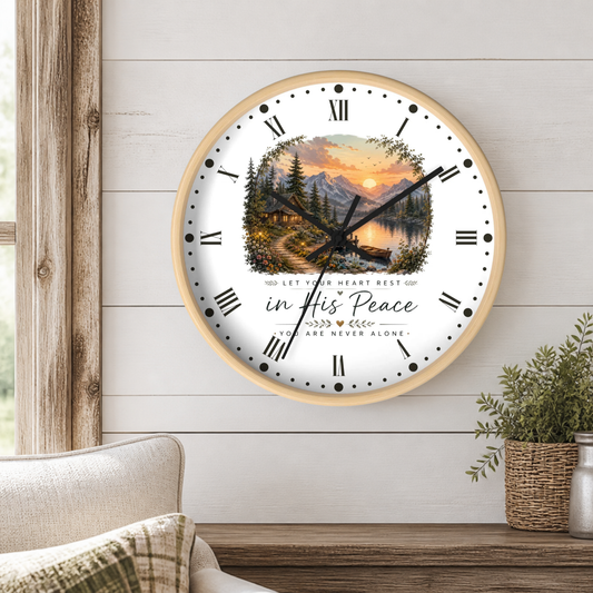 A round wall clock with a light wood frame hangs on a white shiplap wall above a wooden table. The clock face features a scenic landscape illustration of a mountain lake at sunset, Roman numerals, black hands, and the text 'in His Peace' and 'YOU ARE NEVER ALONE.