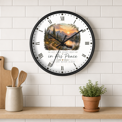 A round wall clock with a black frame and a scenic forest lake sunset print on the face, mounted on a white subway tile wall above a wooden shelf with kitchen utensils and a potted plant.