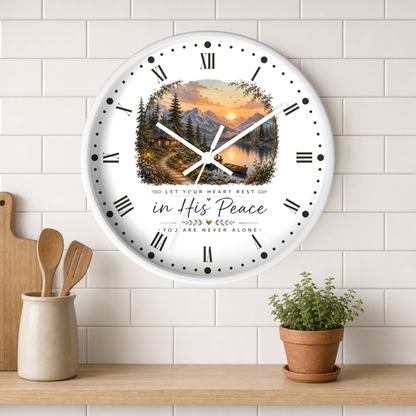 A round wall clock with a scenic mountain and lake sunset print in the center, Roman numerals around the edge, and the words 'in His Peace' and 'You Are Never Alone' below. The clock is mounted on a white subway tile wall above a wooden shelf with a small potted plant and utensils.