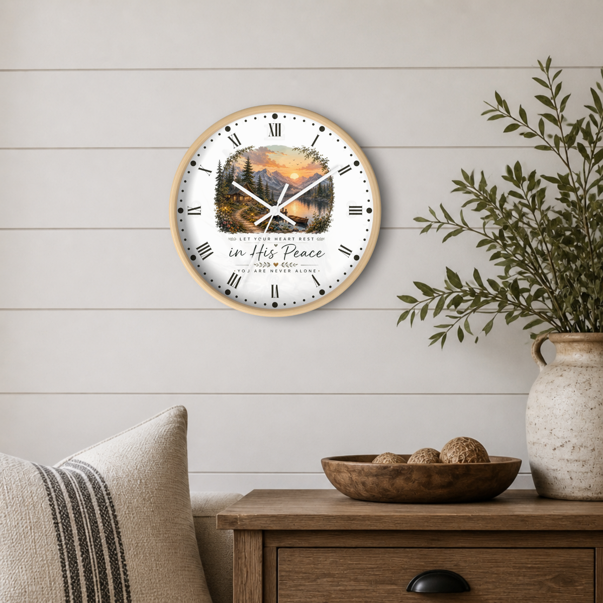 A round wall clock with a light wood frame hangs on a white shiplap wall above a wooden side table. The clock face features a scenic mountain landscape print with a sunset and the text 'In His Peace' and 'God Bless'.