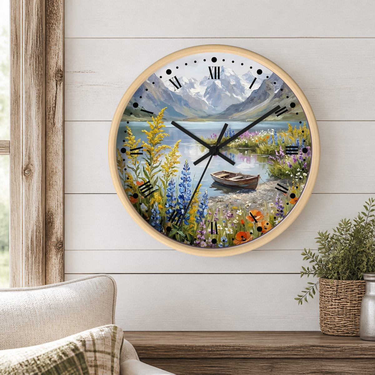 A round wall clock with a light wood frame hangs on a white shiplap wall above a wooden table. The clock face features a colorful landscape illustration of a mountain lake with wildflowers, a small rowboat, and black Roman numerals.
