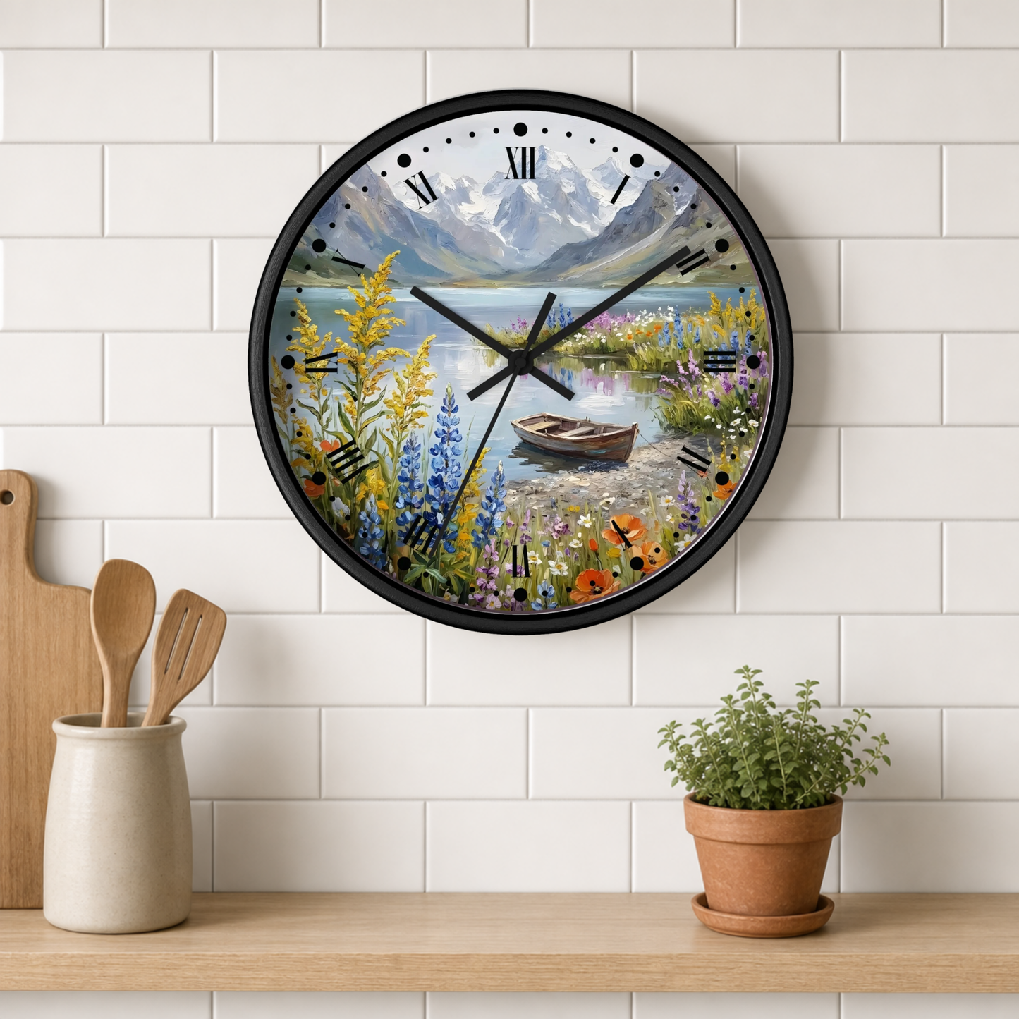 A round wall clock with a black frame and a scenic landscape print of a mountain lake, wildflowers, and a small boat. The clock is mounted on a white subway tile wall above a wooden shelf with kitchen utensils and a potted plant.