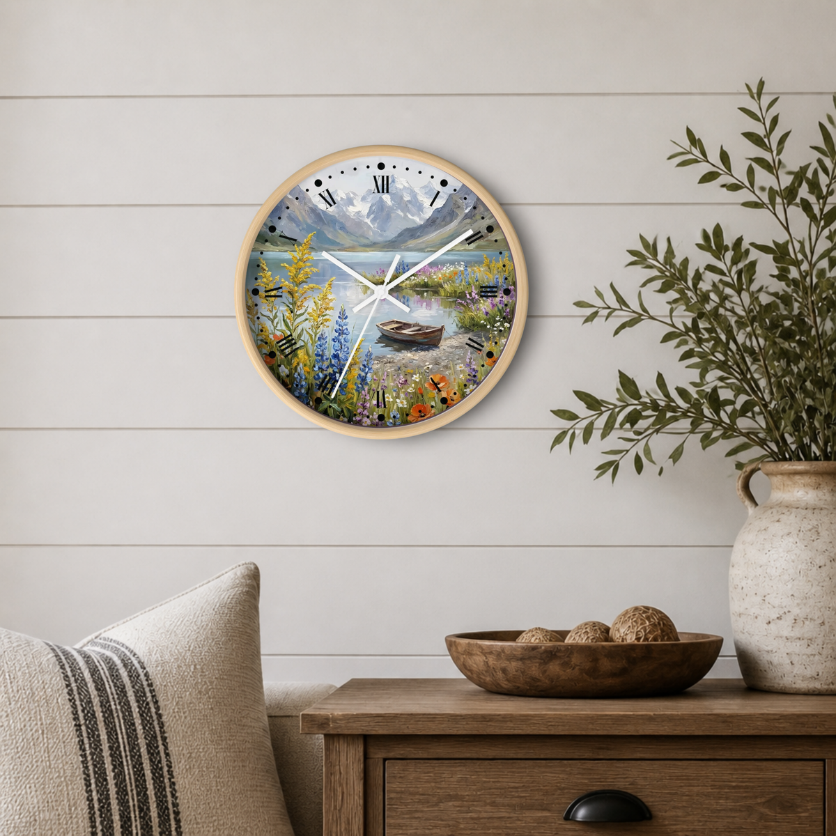 A round wall clock with a light wood frame and a colorful landscape print featuring mountains, a lake, flowers, and a campfire. The clock is mounted on a white shiplap wall above a wooden side table with a bowl, decorative pillows, and a potted plant.