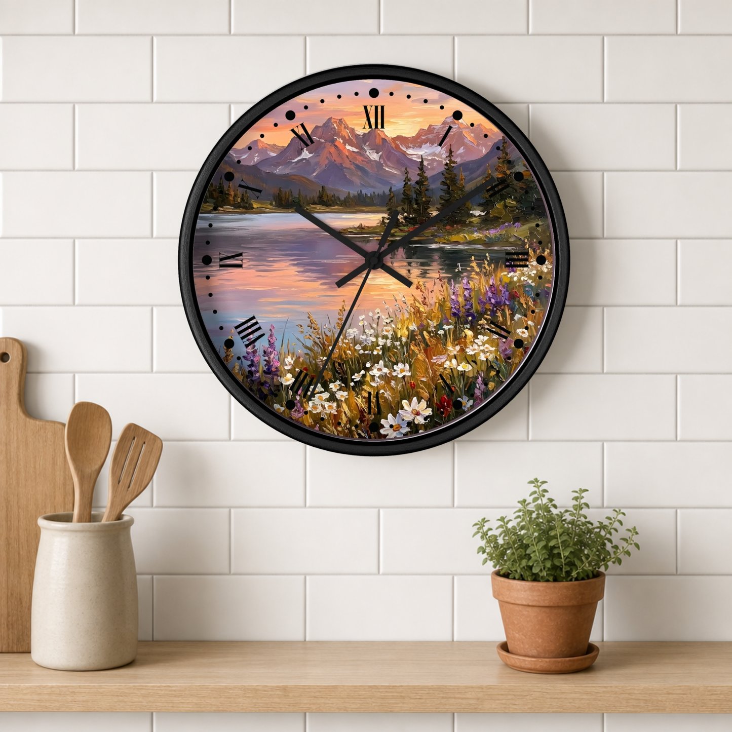 A round wall clock with a black frame and a scenic landscape print of mountains, a lake, and wildflowers, mounted on a white subway tile wall above a wooden shelf with kitchen utensils and a potted plant.