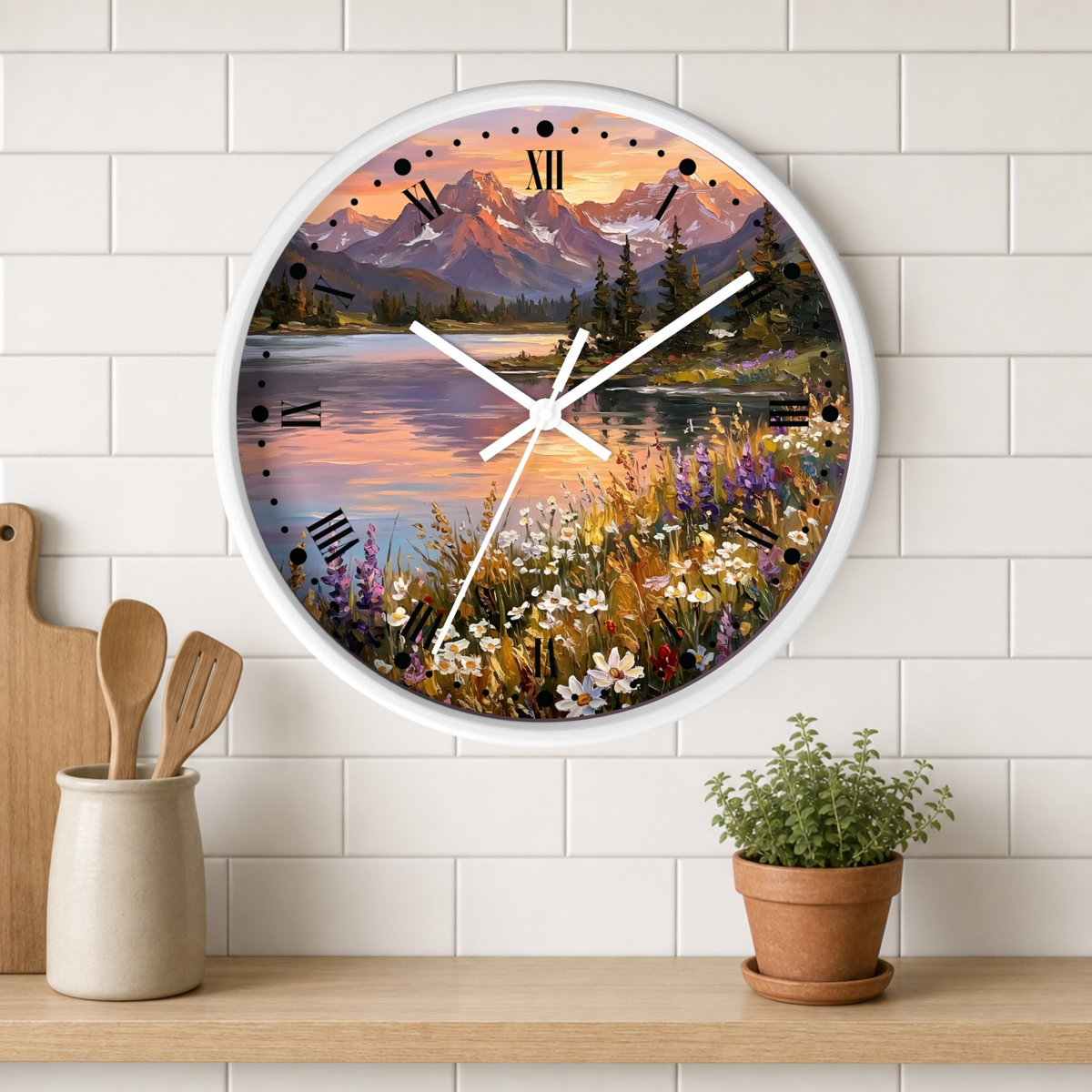 A round wall clock with a white frame hangs on a white subway-tile kitchen wall above a wooden shelf. The clock face features a colorful landscape print of mountains, a lake, and wildflowers at sunset.