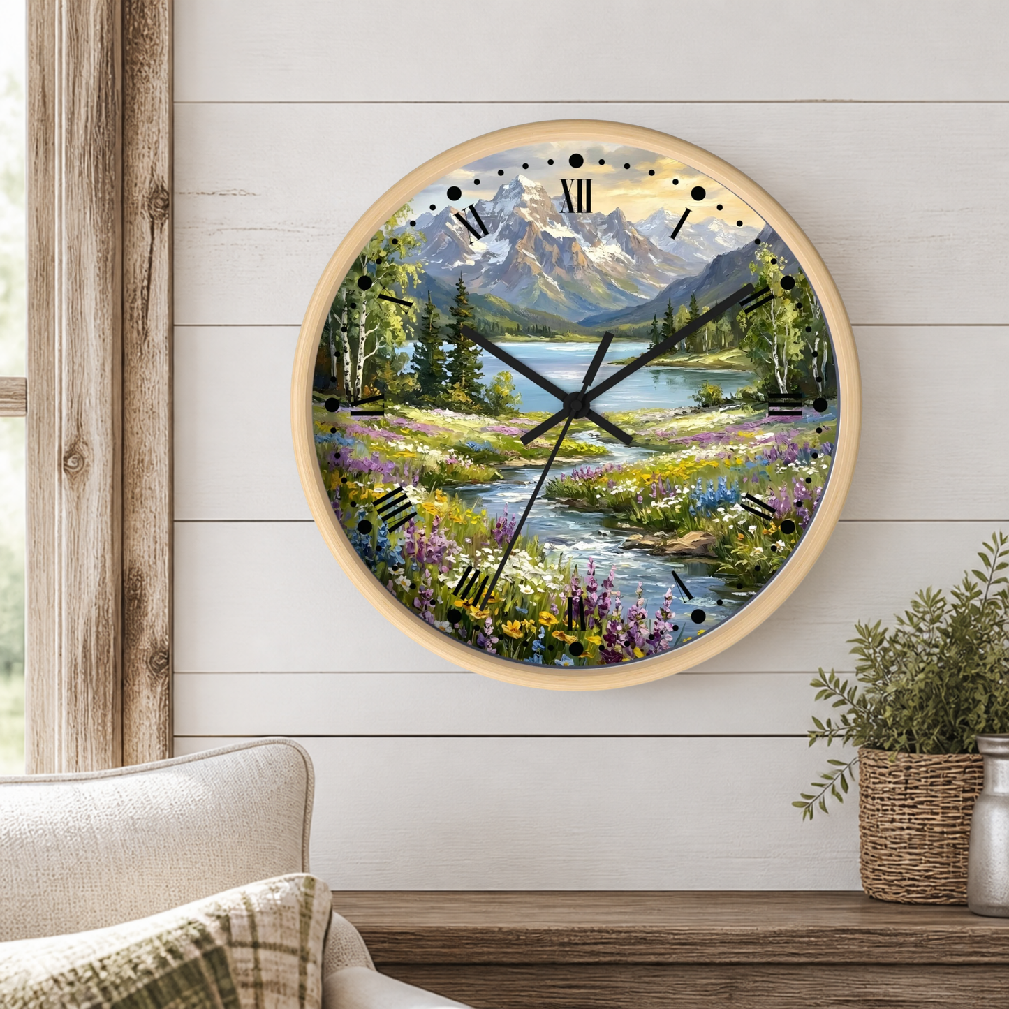 A round wall clock with a light wood frame and a scenic mountain lake landscape printed on the face, mounted on a white shiplap wall above a wooden table with a potted plant.