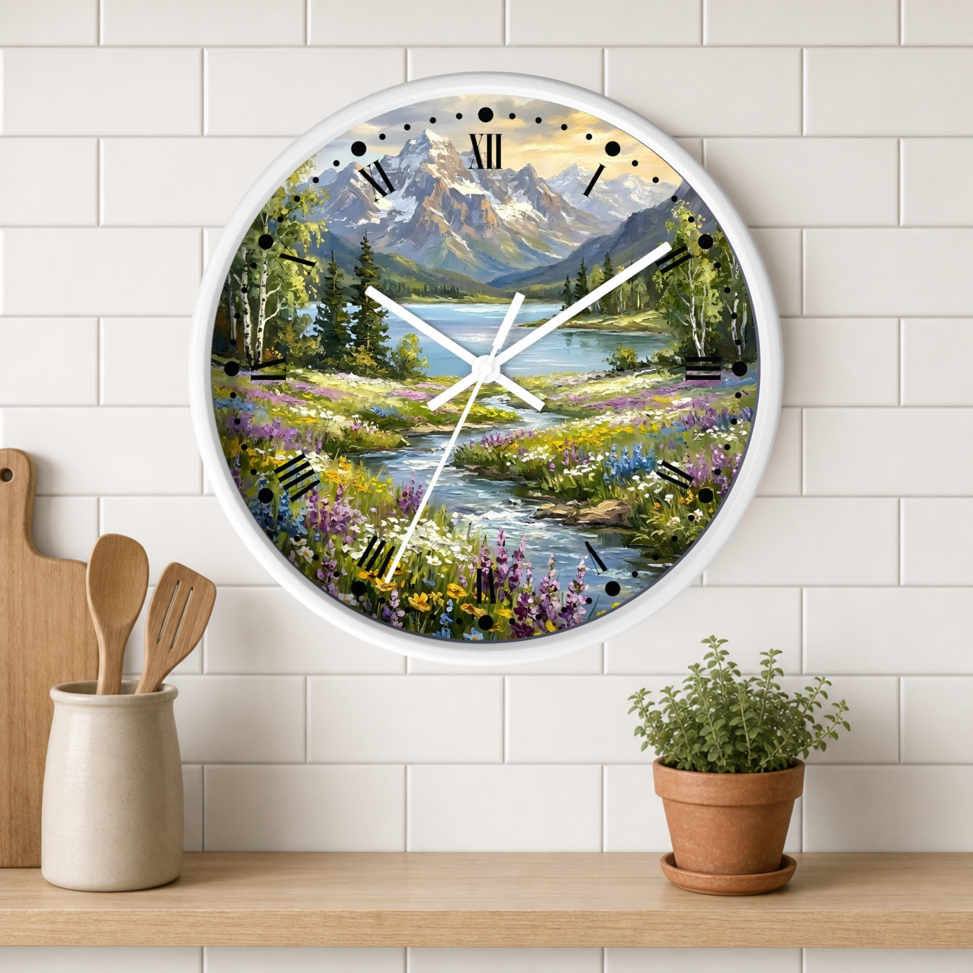 A round wall clock with a white frame and a scenic landscape print of a mountain lake and wildflower meadow, mounted on a white subway tile wall above a wooden shelf with kitchen utensils and a potted plant.