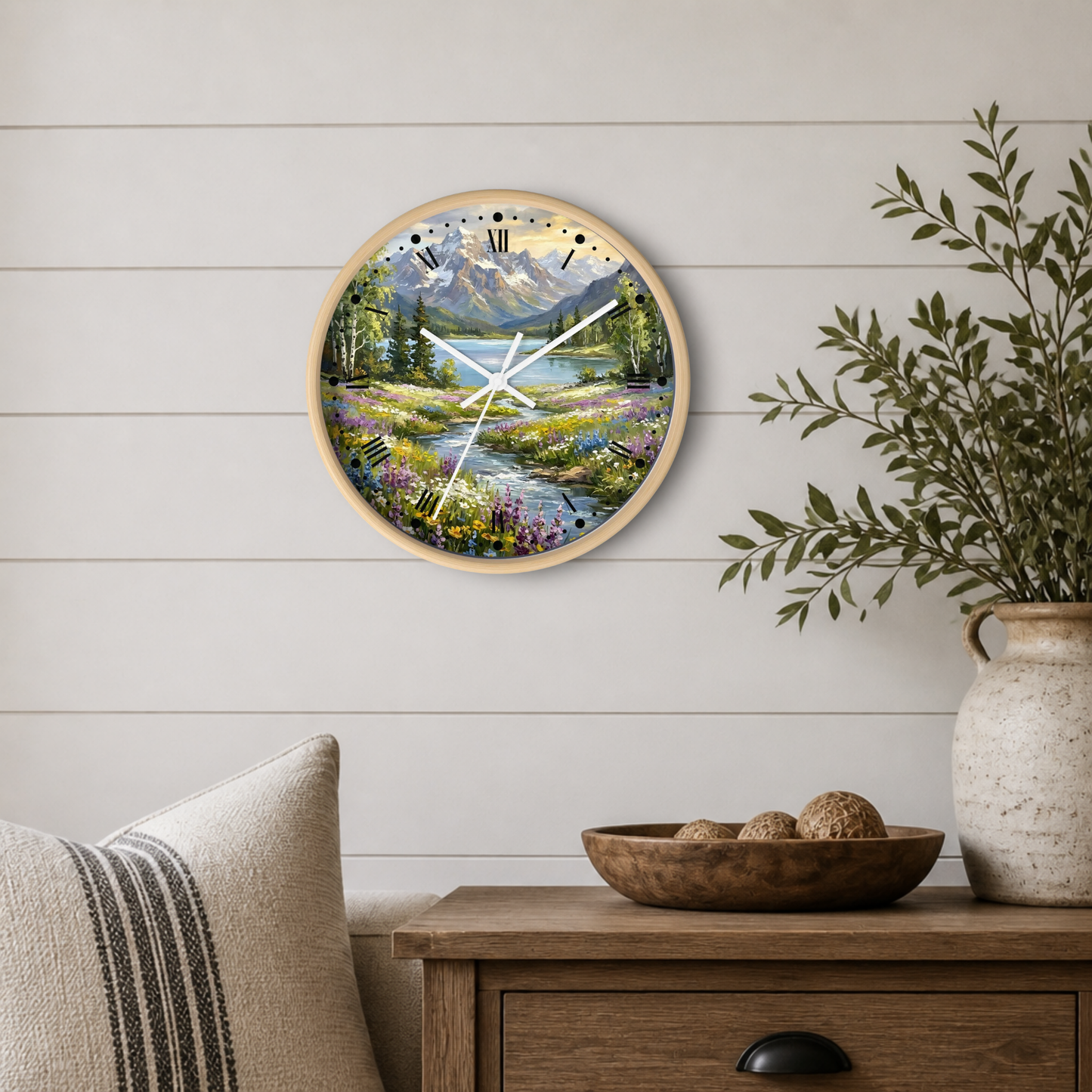 A round wall clock with a natural wood frame and a colorful landscape print of mountains, trees, and a stream, styled on a white shiplap wall above a wooden side table with a vase and pillows.