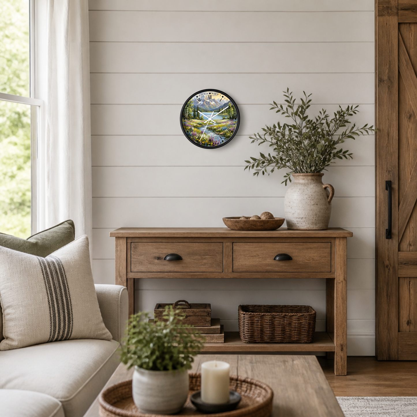 A round wall clock featuring a bright colored mountain lake landscape illustration hangs on a white shiplap wall above a wooden console table in a styled living room setting.