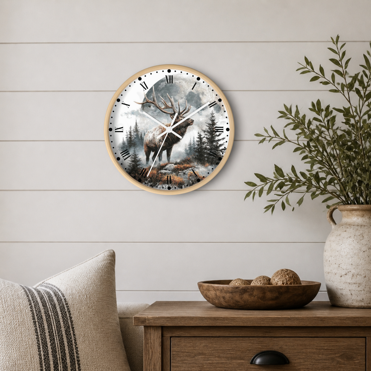 A round wall clock with a light wood frame hangs on a white shiplap wall above a wooden side table. The clock face features a scenic forest illustration with an elk and is styled in a cozy room setting with a vase of greenery and a striped pillow.