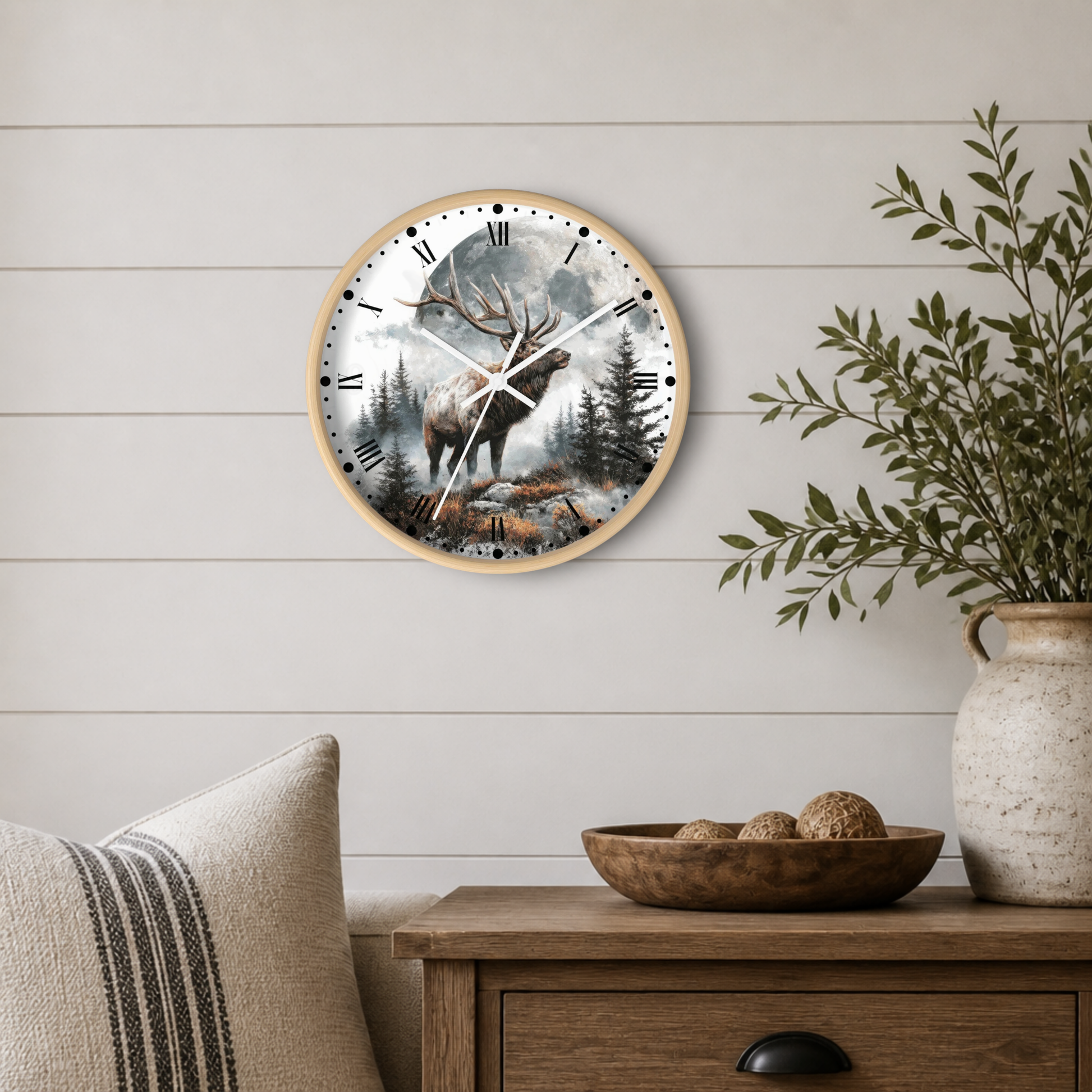 A round wall clock with a light wood frame hangs on a white shiplap wall above a wooden side table. The clock face features a scenic forest illustration with an elk and is styled in a cozy room setting with a vase of greenery and a striped pillow.