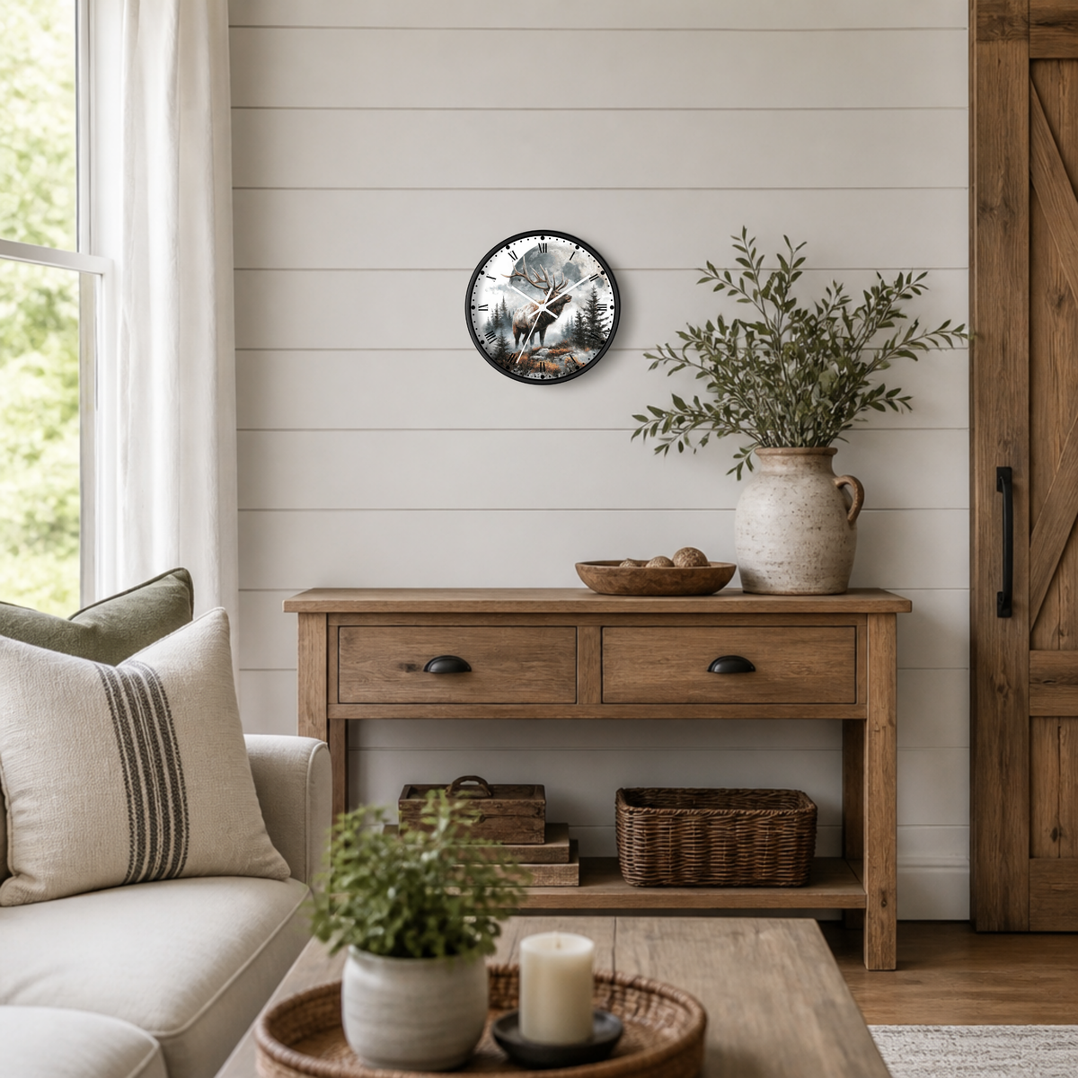 A round wall clock with a black frame and a detailed elk illustration, styled on a white shiplap wall above a rustic wooden console table in a bright, modern farmhouse living room.