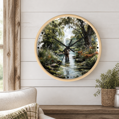 Round wall clock with a light wood frame and a printed scenic dial showing a fly fisherman standing in a creek surrounded by lush green trees and wildflowers. The clock is mounted on a white wood-paneled wall above a wooden shelf with a potted plant and part of a beige armchair visible.