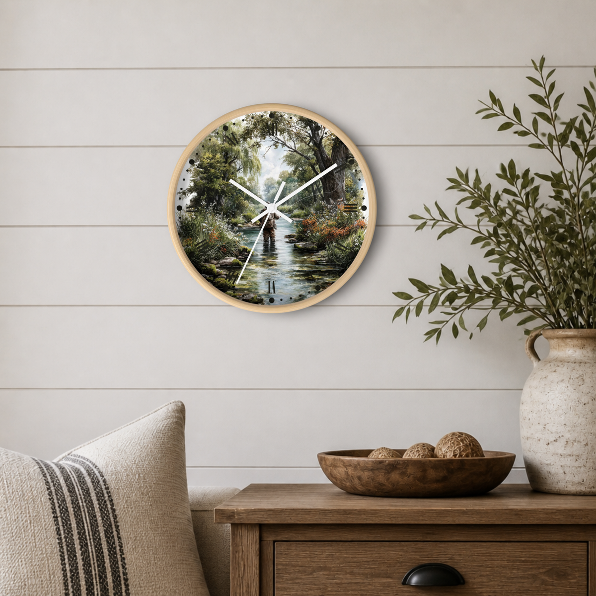 Round wall clock with a light wood frame and a scenic forest stream print, mounted on a white shiplap wall above a wooden side table with a plant and decorative bowl.