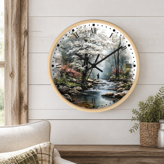 A round wall clock with a light wood frame hangs on a white shiplap wall above a wooden table. The clock face features a scenic forest illustration with a stream, trees, rocks, and a person walking.