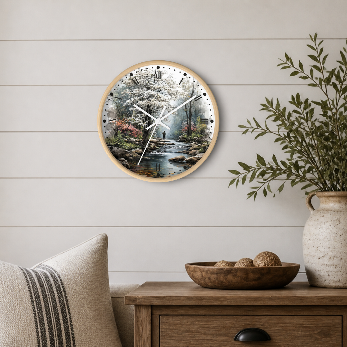 Round wall clock with a light wood frame and a scenic landscape print of a stream and cherry blossoms, styled on a white shiplap wall above a wooden side table with a plant and decorative bowl.