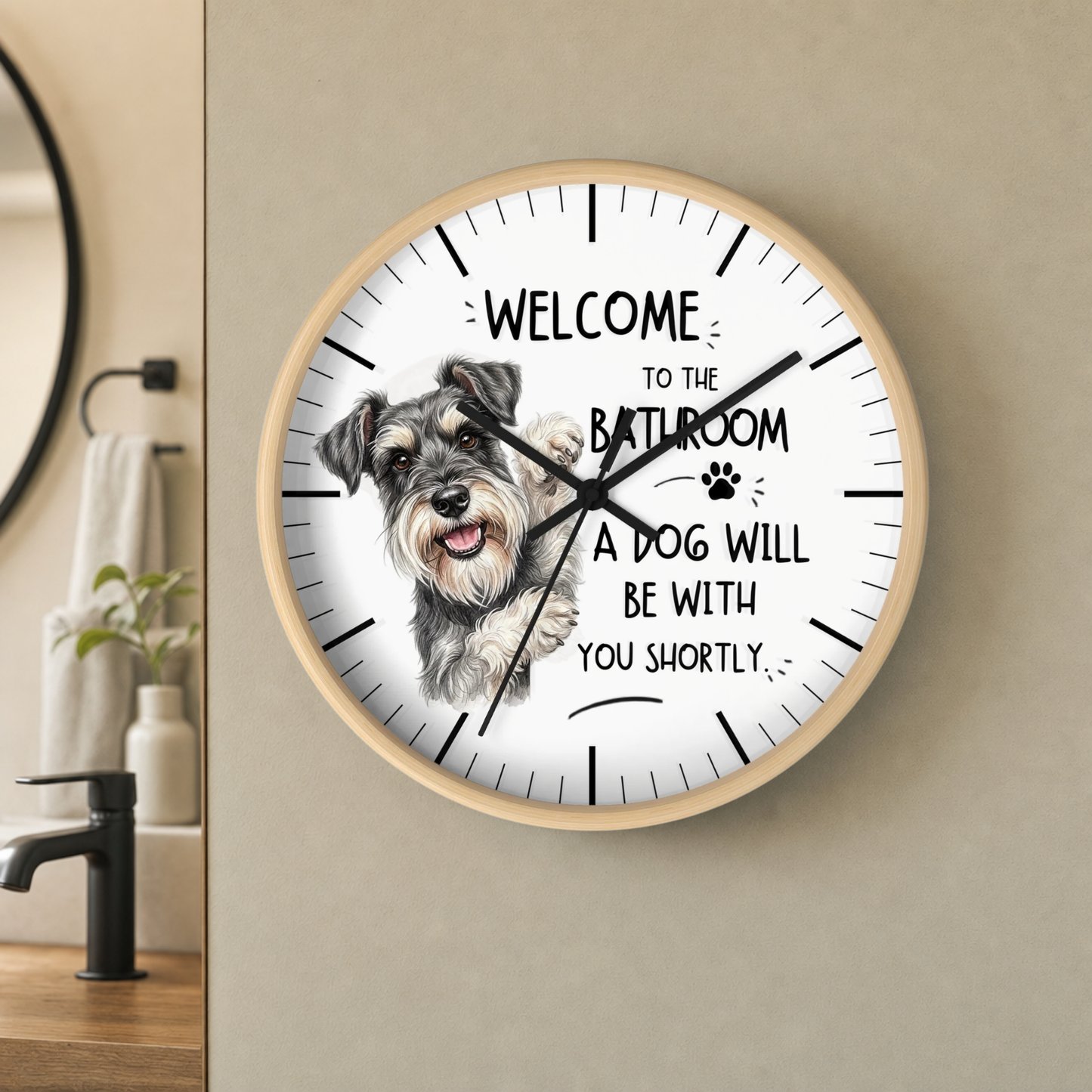 A round wall clock with a light wood frame and a white face, featuring a sketch of a gray and white dog and the text 'WELCOME TO THE BATHROOM A DOG WILL BE WITH YOU SHORTLY'. The clock is mounted on a beige wall in a bathroom setting.