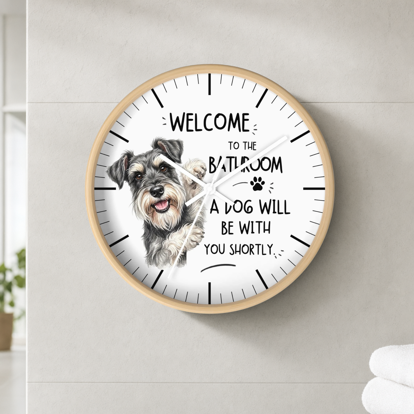 A round wall clock with a light wood frame and a white face, featuring a sketch of a gray and white dog and the text 'WELCOME TO THE BATHROOM A DOG WILL BE WITH YOU SHORTLY'. The clock is mounted on a light gray tiled wall.