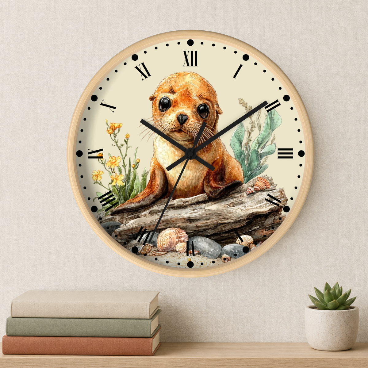 A round wall clock with a light wood frame and a cream dial, featuring a watercolor illustration of a brown seal on a log with rocks and flowers. The clock is mounted on a textured beige wall above a shelf with stacked books and a small potted succulent.