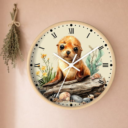 Round wall clock with a watercolor illustration of a seal pup sitting on driftwood, surrounded by seashells, green seaweed, and small yellow flowers on a cream dial with black Roman numerals. The clock is mounted on a warm beige wall with a dried herb sprig hanging to the left.