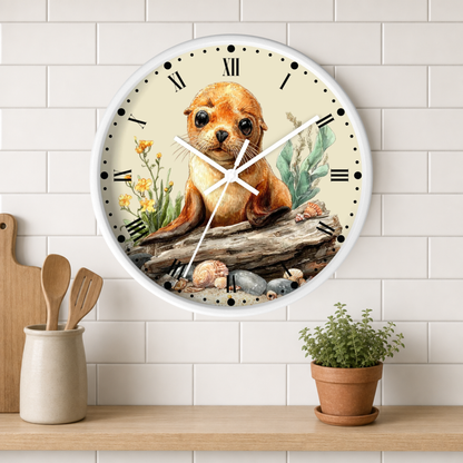 A round wall clock with a white frame and a printed illustration of a brown seal pup sitting on driftwood among rocks and yellow flowers. The clock is mounted on a white subway-tile wall above a wooden shelf with a small potted plant and wooden utensils.